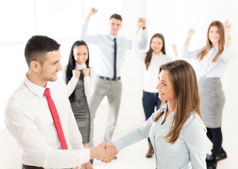 Successful young businesswoman and businessman shaking hands over a deal in front their business team with hands raised celebrating.