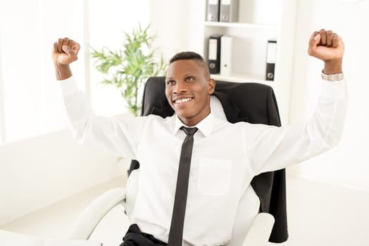 Successful African businessman celebrating success with raised arms in the office. Looking away.