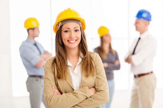 Successful smiling female architect with helmet standing in the office and looking at camera. Her colleagues talking in background.