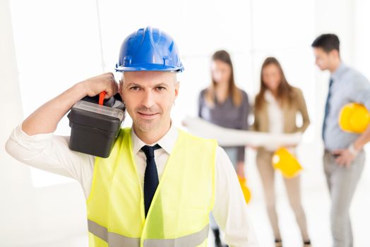 Smiling electrician with helmet holding toolbox in the office and looking at camera. His colleagues talking in background.