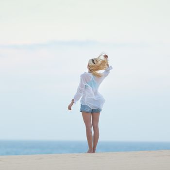 Meditative, sensual blonde woman wearing white loose casual shirt on vacations, looking at horizon, enjoying peaceful early morning.