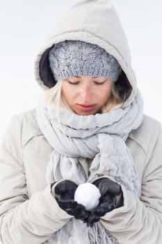 Cute casual young woman wearing glooves, woolen cap and scarf, holding icy snowball in cold winter time. Lady looking down at snowball.