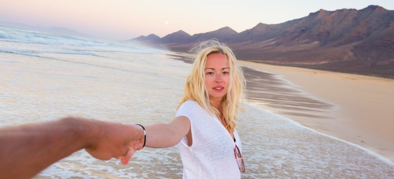 Young romantic couple, holding hands, having fun on perfect deserted beach at sunset. Shot from boyfrieds perspective. Guy looking at her beautiful carefree girlfriend.