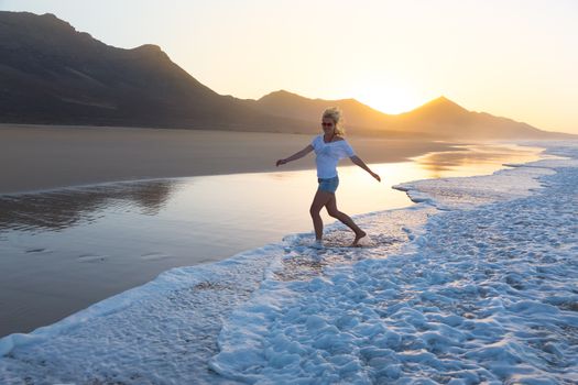 Woman having fun runing from waves on solitary sandy beach in sunset. Waves sweeping away her traces in sand. Beach, travel, concept. Copy space. 