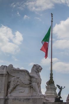 Detail of the Vittorio Emanuele Monument in Italy, Rome, Piazza Venezia. Statue of a lion with wings with an italian flag in the background. Cloudy sky.