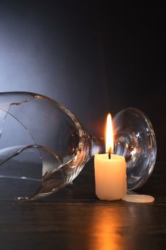 Broken wineglass near lighting candle on dark background