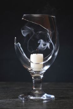 Extinguished candle with smoke inside broken wineglass on dark background