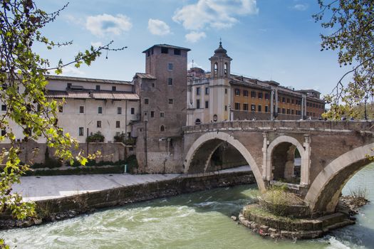 Green water of river Tiber, surrounding the Island with historical buildings and an old bridge. Cloudy blue sky.