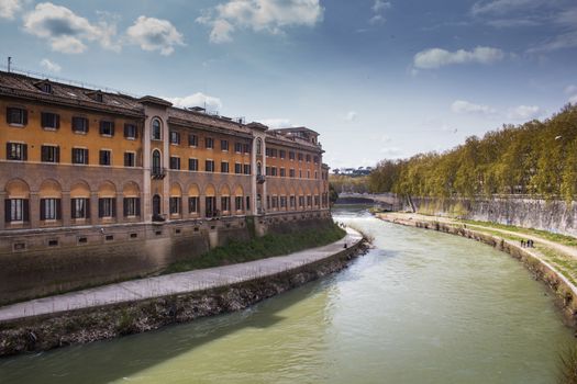 Green water of river Tiber, surrounding the Island with historical buildings. Trees alley on the right riverbank. Cloudy blue sky.