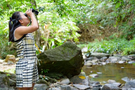 Beautiful young girl hiking is using binoculars look for birds in tropical forest near the waterfalls in Thailand
