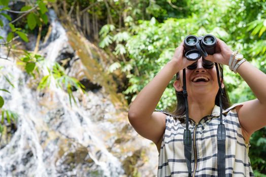 Beautiful young girl hiking is using binoculars look for birds in tropical forest near the waterfalls in Thailand
