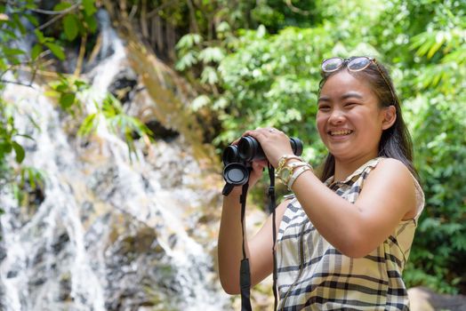 Beautiful young girl hiking is using binoculars look for birds smiling to the camera in tropical forest near the waterfalls in Thailand