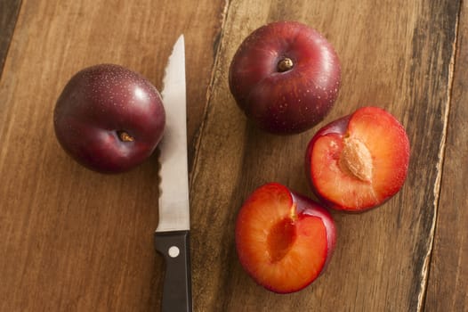 Fresh whole and halved plums showing the stone or pip lying on a wooden table with a knife in a close up overhead view