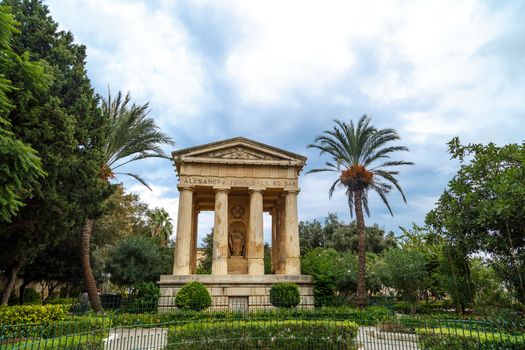 View of Lower Barrakka Gardens with historical monumental columns in Malta, with small trees around, on cloudy blue sky background.