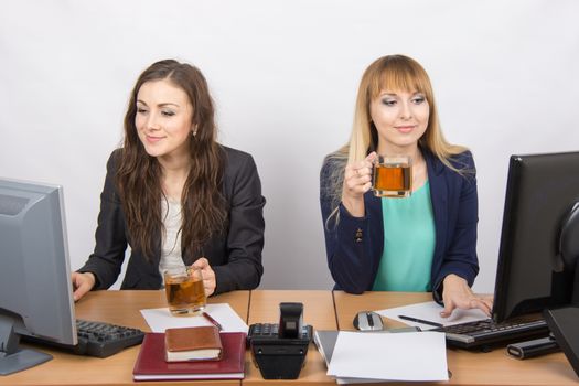 Two young beautiful girls drink tea, office behind a desk and working on the computer