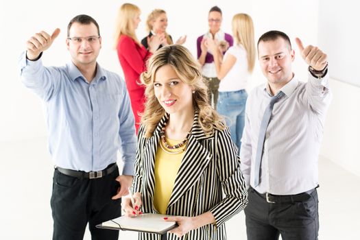Smiling businesswoman standing in the office in front of her colleagues. Her co-workers showing Thumbs Up. They are looking at camera.