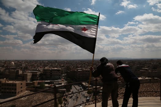 SYRIA, Saqba: Two men hold a pre-Baath Syrian flag, adopted during the uprising in 2011, and watch an anti-regime demonstration in the rebel-held town of Saqba, on the outskirts of the Syrian capital Damascus, on March 18, 2016. Syria's war is entering its sixth year with a glimmer of hope that a landmark ceasefire and a push for peace could help resolve a conflict that has sent hundreds of thousands fleeing to Europe. The conflict began with anti-regime protests in mid-March 2011 and spiralled into a bloody war after a harsh government crackdown on demonstrators.