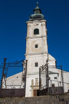 Entrance to the Church of Saint Cross. Old facade, high tower with clock. Cross on the tower. Bright blue sky.