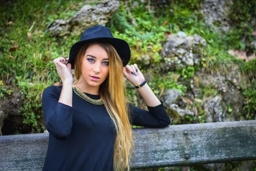 Pretty blonde young woman outdoor in city park, wearing short black dress and fedora hat