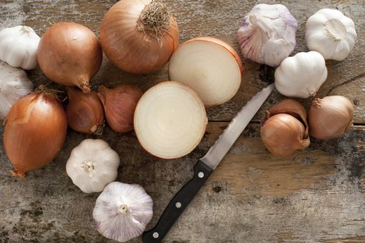 Whole and halved fresh uncooked garlic and onions of the Allium family with a knife on an old rustic kitchen table ready to prepare for cooking