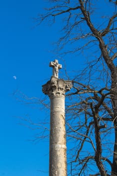 Column with a cross under old big tree. Bright blue sky with a moon. Devin, Bratislava, Slovakia.