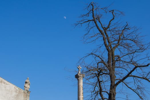 Part of a roof of Church of Saint Cross with a statue. Column with a cross under old big tree. Bright blue sky with a moon. Devin, Bratislava, Slovakia.