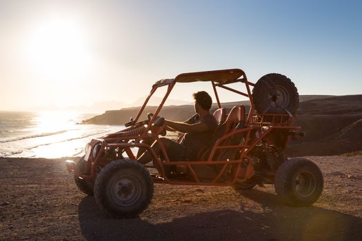 Active man driving quadbike on dirt road by the sea in sunset.