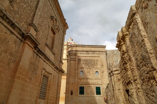 View of Mdina streets with limestone historical buildings around, on cloudy sky background.
