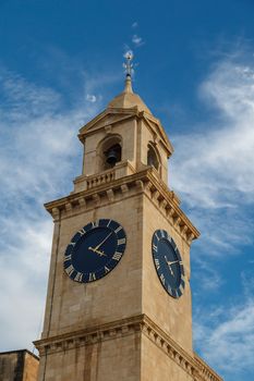 View of a church in Valletta Malta with limestone belfry clock tower, on cloudy blue sky background.