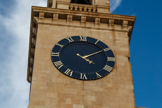 View of a church in Valletta Malta with limestone belfry clock tower, on cloudy blue sky background.