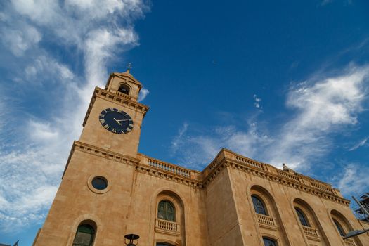 View of a church in Valletta Malta with limestone belfry clock tower, on cloudy blue sky background.
