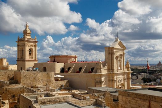 View of a historical church in Victoria, Malta with a limestone belltower, on cloudy sky background.