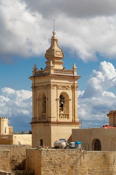 View of a historical church in Victoria, Malta with a limestone belltower, on cloudy sky background.