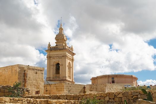 View of a historical church in Victoria, Malta with a limestone belltower, on cloudy sky background.