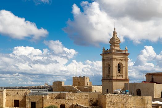 View of a historical church in Victoria, Malta with a limestone belltower, on cloudy sky background.