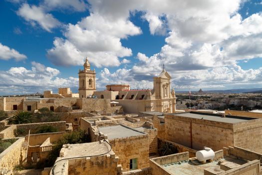View of a historical church in Victoria, Malta with a limestone belltower, on cloudy sky background.