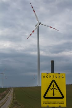 Wind turbine and an early spring field in Berg, Austria. Yellow Attention Board in german language. Rainy cloudy sky.