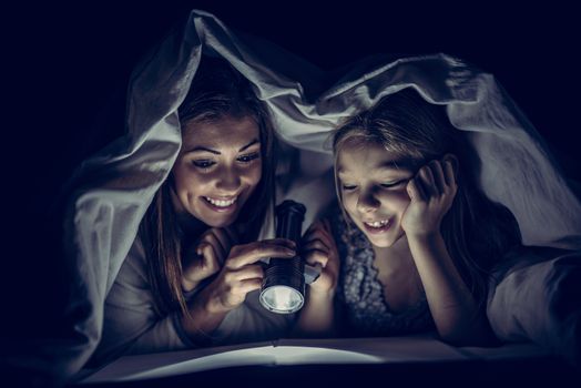 Smiling mother and her daughter reading book under bed cover and holding a flashlight. 
