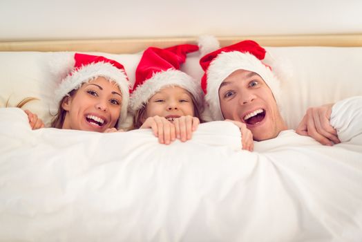 Beautiful smiling family with santa hat having fun in bed. They lie in bed looking at camera and peeking under the blanket with smile.