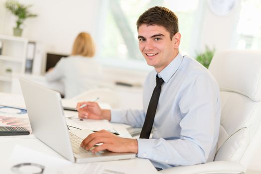 Young businessman sitting at an office desk in front of a laptop and working. Looking at camera.