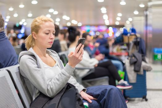 Casual blond young woman using her cell phone while waiting to board a plane at the departure gates. Wireless network hotspot enabling people to access internet conection. Public transport.