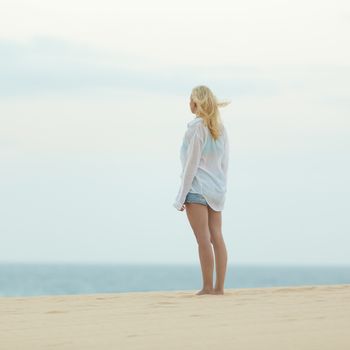 Meditative, sensual blonde woman wearing white loose casual shirt on vacations looking at horizon at dusk.