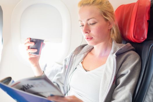 Woman reading magazine and drinking coffeeon airplane. Female traveler reading seated in passanger cabin. Sun shining trough airplane window.