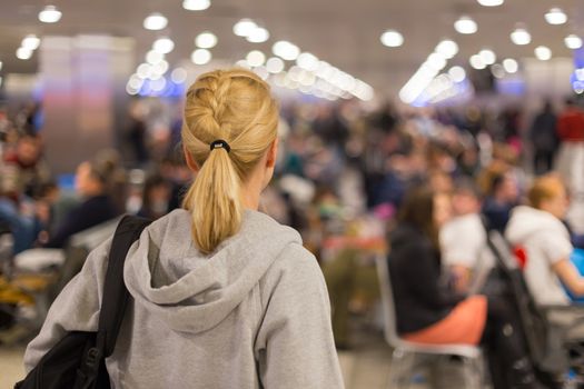 Young blond caucsian woman waiting on airport terminal full of passenegers waiting to depart.