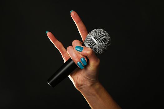 Woman hand with blue nails holding microphone with devil horns rock metal sign isolated on black background