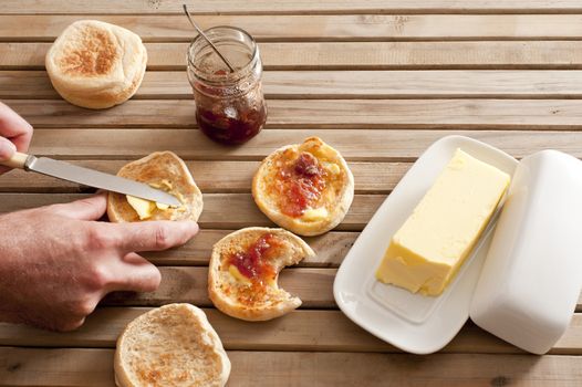 One stick of yellow butter in its serving tray beside a pair of hands spreading jelly on four english muffins with knife