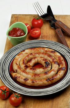 Delicious Grilled Spiral Sausage with Ketchup and Cherry Tomatoes on Striped Plate with Rustic Fork and Knife on Wooden Cutting Board closeup on White Plank background