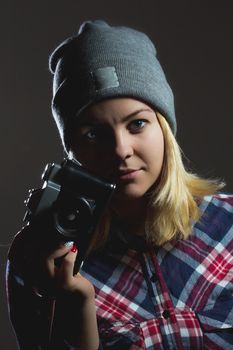 Portrait of hipster girl posing with retro camera wearing checkered shirt and winter hat on dark background