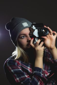 Portrait of hipster girl taking picture with retro camera wearing checkered shirt and winter hat on dark background
