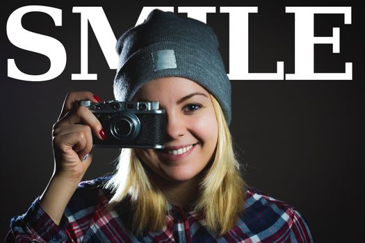 Portrait of hipster girl taking picture with retro camera wearing checkered shirt and winter hat with type SMILE on the background